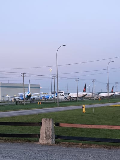 The image depicts an outdoor scene at the Montreal–Mirabel International Airport in Mirabel, Canada. The sky is a pale, dusky blue, suggesting either dawn or dusk. In the mid-ground, several commercial aircraft are parked on the tarmac. Two aircraft with blue and white livery are on the left, with one prominently displayed from the rear, showing its tailfin. To the right, two white Air Canada aircraft are parked, their red maple leaf logos visible on the tailfins. A large, light-colored building, possibly a hangar or terminal, is situated behind the aircraft on the left. Overhead power lines crisscross the scene, supported by wooden utility poles. Streetlights are also visible, some illuminated. In the foreground, a wooden fence with two dark horizontal planks is in sharp focus. Beyond the fence is a grassy area, and further back, a paved road or taxiway. A bright yellow fire hydrant stands on the grass in the middle right of the frame. The ground in the immediate foreground is covered in grey gravel. There are no discernible people or animals in the image.