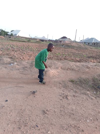 A young boy wearing a green t-shirt and blue jeans with sandals is standing on a dirt path in what appears to be a rural or semi-rural area. Behind him is a field with rows of young plants, likely crops. In the background, there are several houses with corrugated metal roofs, suggesting a residential area. The sky is overcast, and the overall lighting suggests it is daytime. The environment consists of dry, reddish-brown soil and sparse vegetation. The image appears to be taken in Barikin Gidar Kanat, Nigeria.