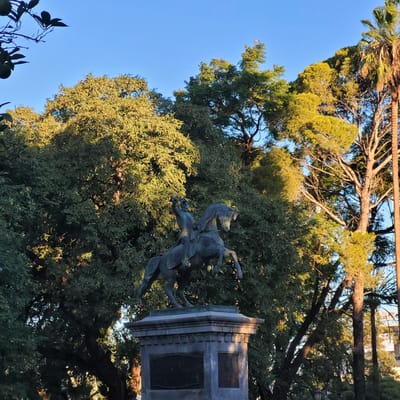 The image shows a bronze equestrian statue on a stone base, set among trees in a park. The statue depicts a rider on horseback, with the horse rearing up. The base has rectangular panels on its sides. The sky is visible in the upper left corner, and parts of buildings are faintly visible through the trees. This statue is located in San Fernando del Valle de Catamarca, Argentina.