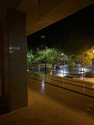 The image depicts a night scene from what appears to be a balcony in Vila Real, Portugal, during or shortly after a rainfall. The balcony has a tiled floor that reflects the streetlights, indicating wetness. A railing provides a partial view of the street below, where parked cars are visible. Trees line the street, their foliage illuminated by the streetlights. In the dark sky, a single streetlight can be seen. The foreground shows a section of the balcony's structure, including a wall or a dividing element. No people are visible.