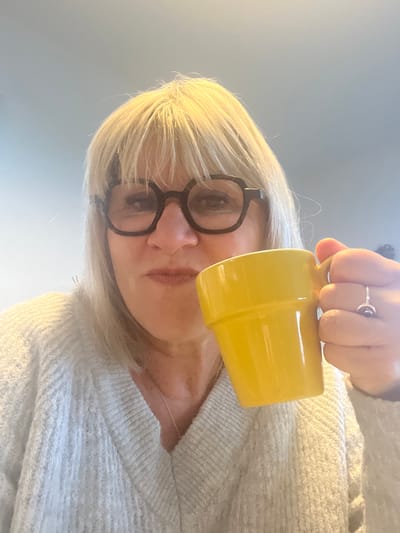 The image, captured in Chambœuf, France, depicts an adult individual in a close-up, frontal composition. The subject has blonde hair with bangs, wears dark, round-framed eyeglasses, and is dressed in a light-colored, V-neck chunky knit sweater. A thin silver chain necklace is visible. The subject's right hand holds a bright yellow, ridged mug to the mouth, indicating an action of drinking or preparation to drink, with lips slightly pursed. A silver ring featuring a dark stone is present on the right ring finger. The background is an indistinct, uniformly light grey or bluish-grey field, lacking specific environmental identifiers.