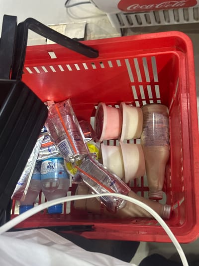A red plastic shopping basket is filled with assorted food and beverage items. These include two glass bottles of milk, several small plastic tubs containing what appears to be yogurt, a package of juice boxes with straws, and some small water bottles. The shopping basket sits in a shopping area, with a cooler for Coca-Cola visible nearby. A portion of a white shirt can be seen in the right foreground of the image.