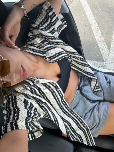 A woman lies on her back in the passenger seat of a car, looking up at the camera. She is wearing a black bikini top, grey sweat shorts, and a black and white striped crochet cardigan. She has on square-shaped sunglasses with gold frames and a silver bracelet on her left wrist. Her fingernails are painted with a light-colored, glittery polish. A tattoo is visible on her left arm. The interior of the car is dark, with black leather seats. Through the open car door, a paved surface with white lines, likely a parking lot or road, is visible, along with some dry leaves and a manhole cover. The lighting suggests it is daytime, possibly sunny, based on the shadows and brightness. The image appears to be a casual, personal photograph taken by the subject herself or someone with her. The location context of Karnobat, Bulgaria is noted but not visually evident in the image itself. There is no visible text in the image.
