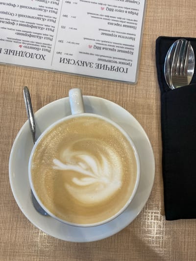 The image shows a top-down view of a table setting. On the table is a cup of coffee with latte art, a white saucer, a spoon, a black napkin with a fork, and a menu in Russian. The menu lists various appetizers, including hot and cold dishes such as chicken wings, nuggets, sushi rolls, and ribs. The table itself has a light brown, textured surface. No people are visible. The location information (Neftekamsk, Russia) is given but not evident from the image's content.