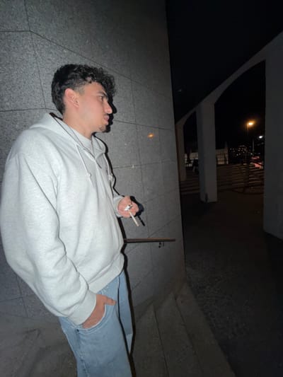 An image taken at night in Vila Real, Portugal, depicts a young adult male in profile, standing and leaning against a textured grey wall. The male subject has dark, curly hair and light facial stubble. He is wearing a light grey zippered hoodie over a lighter undershirt and light blue jeans, with his left hand partially obscured in his left jean pocket. His right hand holds a lit cigarette, from which visible smoke emanates. He is looking towards the right side of the frame. To the right of the subject, the scene transitions to a darker area with architectural elements; two thick, light-colored pillars support an arched overhead structure, framing a darker background. A dark handrail is partially visible in the lower-left. The ground appears to be dark, possibly paved, with visible steps in the mid-right leading upwards. In the distant background, a bright orange streetlamp illuminates a dark urban landscape, indicating an outdoor nighttime setting.
