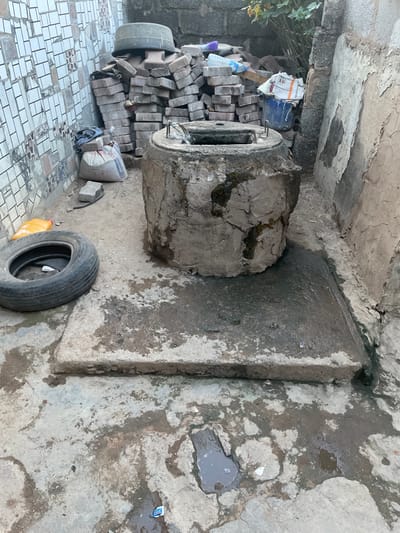 The image displays a cluttered and somewhat neglected outdoor or semi-enclosed utility area, possibly a backyard corner, storage space, or alleyway. The scene is illuminated by natural light, suggesting daytime, without harsh shadows.
In the center of the frame, a large, rough concrete cylinder or pipe segment lies horizontally on the ground. It appears old and weathered, with dark stains and patches of moss or grime clinging to its surface. One end of the cylinder is visible, revealing a hollow interior and what looks like some protruding metal rods or rebar.
To the left of the concrete cylinder, there is a large, irregularly stacked pile of light grey, interlocking pavers or bricks. Some plastic bottles and small pieces of debris are mixed in with the bricks. Behind this pile, a black tire or wheel rim is partially visible, standing upright against a concrete or block wall. Further left and towards the bottom, the ground transitions to an area paved with white and grey square tiles. A white sack, possibly containing cement or other building material, is situated near the base of the brick pile, with the word "BANCUT" clearly visible on its side. Some green foliage from a plant or bush is visible in the upper left corner, growing near the wall.
To the right of the concrete cylinder, a flat, rectangular concrete slab or raised platform extends from a wall. This slab, along with the surrounding ground, is discolored and stained, with several puddles of water indicating dampness. On the lower right side of the foreground, another black tire lies flat on the ground next to a bright yellow plastic jerry can. Scattered across the concrete ground and within the puddles are various small pieces of trash and debris, including wrappers and plastic fragments.
The walls in the background and to the right are made of rough concrete or plastered material, showing signs of wear, cracking, and peeling. An overhead concrete structure or ceiling is also visible at the top of the image, similarly aged and discolored. The overall impression is one of disuse, wear, and a lack of maintenance.