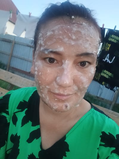 A woman is taking a selfie outdoors. She has a white substance, possibly a face mask, on her face and forehead. She is wearing a green and black floral patterned top. In the background, laundry is hanging on a clothesline, and a fence is visible. A sign with text, including ".AJ" and some reversed writing, is hanging on another clothesline. The scene appears to be in a residential area.