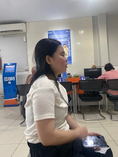 An outdoor image shows a woman sitting and holding a smartphone. The woman has long, dark hair and is wearing a white top and black pants. She is looking down at her phone, and her face is partially obscured by her hair. The phone screen displays an image of a light-colored object against a dark background. In the background, there is a bank, a BRI ATM machine, and other people sitting in chairs. The overall scene suggests a waiting area at a bank in Ta', Indonesia.