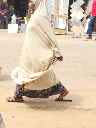 The scene is an outdoor street in Maiburiji, Nigeria. A woman in a light cream-colored hijab and patterned pants stands on a dusty sidewalk. She is wearing sandals. Nearby, several tiles are displayed on a stand, some with varying patterns and colors. Other people are visible in the background, including someone wearing a red shirt and jeans and someone sitting on a chair wearing a striped shirt. There are various objects such as tiles, wooden crates, and plastic containers. The overall setting suggests a marketplace or roadside shop.