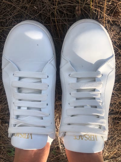A top-down, close-up shot shows a pair of white leather Versace sneakers with white laces. The sneakers are positioned on a bed of dry, brown pine needles, suggesting an outdoor environment. The left sneaker displays the "VERSACE" logo in gold lettering on its heel. The lighting suggests it is daytime, with a sunbeam casting a bright highlight on the toe of the left shoe. The background is a blur of pine needles and some twigs, indicating a natural setting, possibly a park or forest area. The image was captured in Rawai, Thailand.