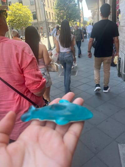 A person's hand in the foreground holds a small, teal-colored gummy candy shaped like a shark. Behind the hand, a person wearing a bright pink shirt walks away from the camera, their arm partially visible. Further in the background, several other individuals are walking down a city street. One woman with long dark hair wears a strapless top and denim shorts, and another man in a black t-shirt and khaki pants is also walking away. The street appears to be paved with dark square tiles and is lined with buildings on the right. Trees with green foliage are visible on the left side of the street, suggesting an outdoor, daytime setting. The architecture and urban environment suggest the location is in Salerno, Italy.