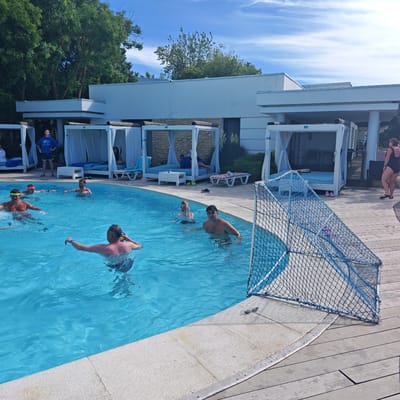 The image captures a lively scene at an outdoor swimming pool on a sunny day. The pool is filled with clear blue water, and several people are enjoying themselves. In the foreground, a man in the pool holds a yellow and blue ball, looking towards the right of the frame. To his left, another man with a yellow headband is partially submerged in the water, with ripples indicating movement. In the background, several white cabana-like structures with draped curtains line the edge of the pool, suggesting a resort or hotel setting. Some people are relaxing in these cabanas, while others are visible in the pool. A man in a blue hooded sweatshirt stands near the cabanas on the left, looking up. The overall atmosphere appears relaxed and recreational. The presence of a blue soccer goal net near the edge of the pool suggests a game or activity is underway or about to begin. The sky is blue with scattered white clouds, indicating fair weather. The lighting suggests it is daytime, likely mid-afternoon. The location context provided is "городское поселение Щелкино, Ukraine," placing the scene in a specific urban settlement in Ukraine.