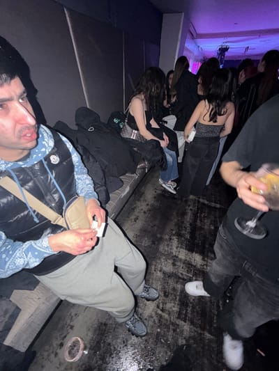 This image depicts an indoor scene within a dimly lit entertainment venue in Vila Real, Portugal, suggestive of a nightclub or bar. The environment features dark, textured walls, a long, dark upholstered bench along the left side, and a dark, worn floor with wet-looking patches and reflections. Overhead, a purple ambient light source illuminates the background.
In the immediate foreground-left, a male with dark hair and a mustache is seated on the bench, looking to the left with a focused expression. He wears a light blue bandana-patterned hoodie under a dark puffer vest (displaying a Moncler logo), light grey sweatpants, and dark athletic shoes. A beige cross-body bag is slung over his shoulder. He holds a small, white rectangular piece of paper and a small, white cylindrical object in his hands, appearing to be preparing a cigarette or similar item.
To the immediate foreground-right, a person's lower torso, arm, and hand are visible. This individual, wearing a dark t-shirt and dark pants, holds a stemmed glass containing a yellowish beverage with ice and a citrus slice. White sneakers are visible on their feet.
In the midground, several individuals are present. Two women are seated on the bench next to the foreground male. One has long dark hair, wears a dark top, light jeans, and white sneakers. A black jacket and a light-colored handbag with a green strap rest on the bench beside her. Another woman is seated further back. In the background, multiple indistinct figures, predominantly women, are seen standing and seated under the purple lighting, indicating a social gathering. On the floor near the foreground male's feet, a small, round ashtray containing dark residue is visible. The exact venue within Vila Real is not discernible from the image.