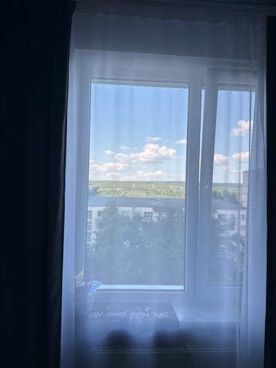 The image shows a view through a partially open window, looking out onto a bright, sunny day. The window is framed by sheer white curtains on the left and a dark curtain at the bottom. The exterior scene reveals a blue sky with scattered white clouds, a strip of green landscape, and several buildings with white facades. Trees are visible in the mid-ground, and the overall impression is one of a suburban or rural setting under a clear sky. The light suggests it is daytime, likely late morning or early afternoon. On the windowsill, near the right side of the frame, a few objects are visible, including what appears to be a box with some writing on it. The text on the box is partially obscured by glare and dust, but some words are discernible, including "Take some Mor". The setting appears to be in Strășeni, Moldova. The mood is peaceful and serene, evoked by the natural light and the quiet outdoor scene.