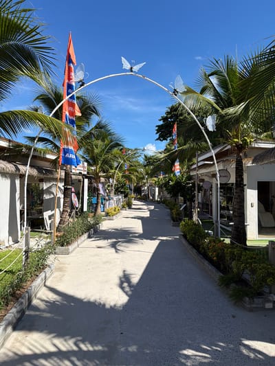 This media file captures a sunny day scene in Baan Chaweng, Thailand. The image showcases a pathway lined with palm trees and small shops, indicative of a tropical resort or commercial area. The pathway itself is made of concrete and is partially shaded by the fronds of palm trees.
Several shops with white facades are visible along the right side of the pathway. One shop has a sign that reads "Beauty," and another sign below it appears to say "Pink Lady." Further down the path, more shops are visible, some with merchandise displayed outside.
Decorative elements are present, including a white archway adorned with translucent butterfly cutouts, suggesting a festive or welcoming atmosphere. Orange and blue flags with white lettering are also visible, adding color to the scene. One flag is particularly prominent, featuring a design that seems to include text, though it's difficult to read clearly.
The overall scene suggests a leisurely environment, likely a tourist destination given the presence of shops and the tropical vegetation. The bright sunlight and clear blue sky indicate a pleasant day, likely mid-morning or afternoon. No people are clearly depicted in the image, allowing the focus to remain on the setting and its decorative elements. The presence of coconuts on one of the palm trees further reinforces the tropical context of Baan Chaweng.