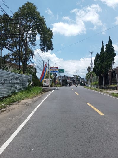 The scene depicts a daytime, wide-angle view of a two-lane asphalt road receding into the distance under a partly cloudy blue sky in Bagochen Boolingpang Ueling, Bhutan. The road features a solid white line marking the left shoulder and broken yellow lines indicating the center.
To the left of the road, a narrow dirt and grass verge runs alongside a corrugated metal fence, covered in dark graffiti. Behind the fence, a dense network of utility poles and black electrical wires is visible, intertwined with a large deciduous tree bearing green foliage. Further down the road on the left, a small white structure, possibly a vendor booth, is present, flanked by vertical red, blue, and white banners or flags. Above these, an "Indomaret" sign is mounted on a pole, and additional low-rise buildings are visible in the background.
To the right of the road, a low stone or concrete fence with decorative pillars separates the roadside from a row of tall, dark green coniferous trees. Beyond these trees, a metallic cellular tower stands against the sky. Further down the road on the right, more buildings are discernible.
A single individual riding a dark-colored motorcycle or scooter is observed in the distant center of the road, traveling away from the camera. No other persons or active vehicles are clearly visible. The overall environment suggests a semi-urban or rural thoroughfare.