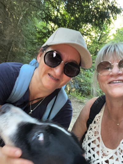 This is a selfie taken outdoors in a wooded area, likely in the commune of Gumières, France, given the context. Two women are in the foreground, with a dog's head partially visible on the left. The woman on the left is wearing a blue t-shirt, a light-colored baseball cap, and large, dark-rimmed sunglasses. She has a light blue backpack on. The woman on the right has blonde hair, is wearing sunglasses with thin, round frames, and a white crocheted top. She is wearing a necklace. The dog is black and white. The background consists of trees and foliage, suggesting a natural, outdoor setting. The lighting indicates it is daytime.