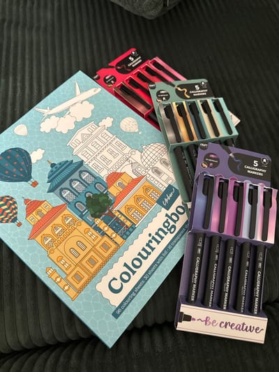 The image shows a collection of art supplies, including several sets of calligraphy markers and a coloring book. The calligraphy markers are packaged in cardboard sleeves, with some sets featuring metallic colors. The coloring book has a light blue cover with an illustration of a city skyline and hot air balloons. The text on the coloring book indicates it contains 96 coloring sheets, with 30 sheets featuring foil and 16 being transparent. The phrase "be creative" is also prominently displayed on one of the marker packages. The scene is set on a dark, textured surface, possibly a sofa or blanket. The lighting is soft and diffused, suggesting an indoor setting. There are no people visible in the image, and the focus is entirely on the art supplies. The overall impression is one of artistic potential and creative engagement. The location context of Stuttgart, Germany, is not directly evident from the image itself, but serves as additional information.