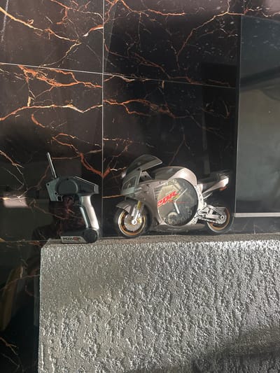 In Brca, Montenegro, on a textured silver shelf, a radio-controlled motorcycle clock sits next to its remote control. The motorcycle, a grey replica with gold accents, features a clock face embedded in its side. The remote control, also grey and silver, has an antenna. Behind the shelf, a black marble-patterned wall with thin gold veining serves as the backdrop. The lighting casts shadows and highlights the reflective surfaces.