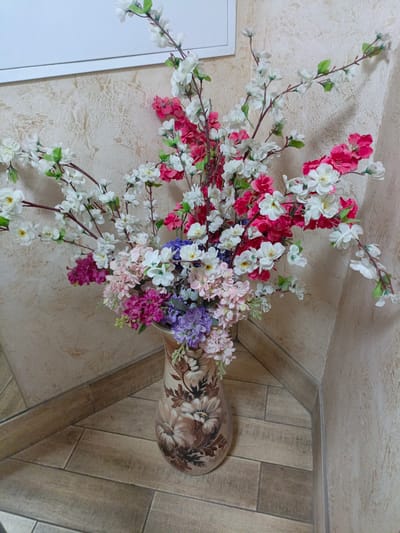 In Sim, Russia, a tall ceramic vase decorated with floral patterns stands on a tiled floor, holding a large bouquet of artificial flowers. The flowers are a mix of white, pink, and purple blossoms, with some green leaves interspersed. The vase is positioned in a corner where two walls meet, with one wall having a textured, light-colored finish and the other appearing to be a lighter, smoother surface. The floor is made of wooden-patterned tiles. The scene is brightly lit, with shadows cast on the wall behind the vase, suggesting a light source from the front. The overall impression is a still-life arrangement in an indoor setting.