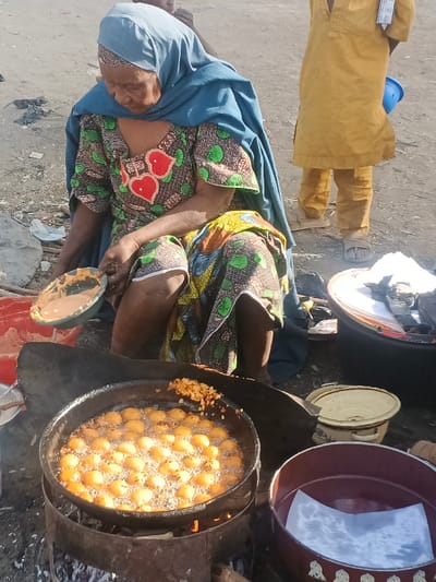 An outdoor scene in Gobirawa, Nigeria, depicts an elderly woman frying food. She is positioned low to the ground, wearing a blue head covering and a patterned dress featuring red hearts and green leaves. Her right hand holds a green bowl containing a light-colored batter, from which she appears to be depositing portions into a large, dark frying pan. The pan rests on a makeshift metal drum over an open fire, actively frying numerous spherical, golden-brown food items in oil; smaller, irregular fried pieces are also visible on the pan's edge. To the right of the cooking setup, a light-colored enamel pot with a lid and a dark reddish-brown plastic basin containing white paper are present. In the background, a person wearing a light brown garment and sandals stands to the right, holding a small blue bucket and with items hanging from their neck. Another individual's head is partially visible behind the cooking woman. The ground consists of unpaved earth with scattered debris.