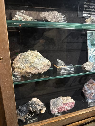 This image shows a display case with several geological specimens. The specimens are labeled with their names and origins. In the foreground, there is a dark, metallic-looking mineral specimen with a label that reads "MALACHIT, MIXIT, BISMUTIT, Alter Schmiedestollen, WITTICHEN". To the right of this, there is a pinkish-red mineral specimen labeled "GED. KUPFER, Malachit, Prosper-Gang, BAD RIPPOLDSAU". Above these, a large, light-colored rock specimen with dark veins is labeled "EMPLEKTIT, Grube Daniel, WITTICHEN". Further up, there are more specimens, including one labeled "GED. SILBER, BLEIGLANZ, Grube Wenzel, OBERWOLFACH". A black sign in the upper right corner reads "DIE FUNDORTE UND HEIMAT GABEN DIE SELTENEN MINERAL. IHRE INTERNATIONAL GOLTIGEN NAMEN". The display case is made of wood and glass. The lighting is dim, with some reflections on the glass. The image appears to be taken in a museum or exhibition. Schonach im Schwarzwald, Germany, is mentioned in the context of the origin of some of the minerals, specifically "WITTICHEN", which is a locality associated with mining in that region.