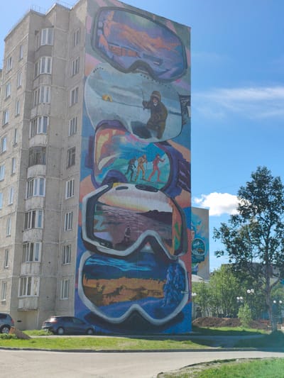 The image shows a mural painted on the side of a multi-story apartment building in Kirovsk, Russia. The mural depicts a series of oversized ski goggles, with different scenes visible through the lenses. The top lens shows a person in a snow-covered landscape, possibly skiing or snowboarding. The second lens from the top features a person ice fishing. The third lens shows three skiers in motion. The fourth lens from the top displays a sunset or sunrise over a body of water, with a lone figure in the foreground. The bottom lens shows a snow-covered landscape with a blue, icy sheen. The building itself is a concrete structure with multiple windows and balconies. In front of the building, there is a grassy area with a road and two parked cars. A large tree stands to the right of the building, and other buildings and trees are visible in the background. The sky is blue with some scattered clouds. The overall scene suggests a depiction of winter sports and arctic landscapes, fitting for a city like Kirovsk, which is known for its proximity to the Khibiny Mountains and winter tourism.
