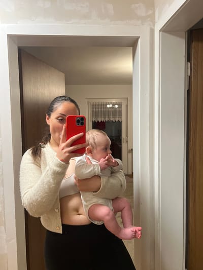 This image depicts an indoor mirror selfie taken by an adult female holding an infant, stated to be located in Wüstenrot, Germany.

The adult female, positioned centrally, has dark hair pulled back and is wearing a cream-colored, fuzzy, open cardigan over a light-colored bra top and dark pants. She is holding a red smartphone with her right hand, positioned to capture her reflection and that of the infant she holds in her left arm. A small red bracelet is visible on her left wrist. Her right eye is partially visible, looking towards the reflected camera.

The infant, held by the adult's left arm, wears a white long-sleeved bodysuit. Its head is turned towards the left of the frame (away from the mirror's direct reflection), and its left hand is near its mouth. The infant's bare feet are visible below the adult's arm.

The background shows an interior space with light-colored walls and white doorframes. To the left of the adult's reflection, an open doorway reveals a room with a window or glass-paneled door draped with white lace curtains, and a light fixture is visible above. To the right, a partially open wooden door is visible through another white-framed doorway. The upper wall section above the doorways displays irregular patches of a lighter shade, indicating unfinished or repaired plasterwork. The lighting appears artificial and even.