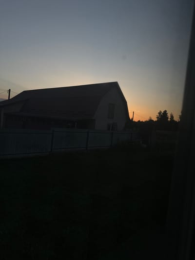 This media file shows a barn with a red roof and white siding, partially silhouetted against a pale orange and gray sky at dusk. The barn appears to be a long, rectangular structure with several windows. To the right of the barn, there is a dark, grassy area, likely a field or lawn. Trees are visible in the distance, also silhouetted against the sky. A utility pole with wires is situated in front of the barn. The overall scene suggests a rural setting at sunset or twilight. There is no visible text in the image.