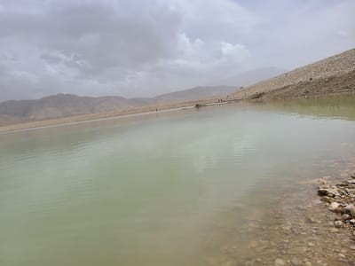 The image displays a large body of water, likely a reservoir or dam, situated within an arid, mountainous landscape near Quetta, Pakistan. The sky is overcast with a continuous layer of grey clouds.
The foreground and mid-ground are dominated by the pale green-beige water, which exhibits subtle surface ripples. In the bottom right corner, a shallow, rocky shoreline is visible, composed of various sizes of brown, grey, and tan stones, partially submerged.
The water body is bordered by engineered embankments on both sides. The embankment on the left, constructed from tightly packed light brown gravel or stone, extends horizontally into the distance, meeting a range of hazy, arid mountains. A single, slender, dark vertical post is partially submerged in the water near this left embankment, centrally positioned in the mid-ground.
The right embankment, similarly constructed from light brown gravel, slopes steeply upwards from the water. On this right embankment, three small, indistinct human figures are observed standing near the waterline in the mid-ground. Further up and to the right on the same slope, a white vehicle, consistent with an SUV or sedan, is parked. A dark, irregularly shaped object is partially submerged in the water, located between the vertical post and the human figures.
The distant background consists of multiple layers of barren, light brown mountains, appearing muted and hazy under the cloudy sky.