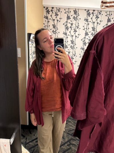 A woman is taking a mirror selfie in a hotel room. She is wearing a rust-colored top and a burgundy open-front shirt over it, along with tan pants. She is holding a smartphone in her right hand and making a kissing face towards the mirror. The background shows a wall with a floral pattern wallpaper and a dark-colored door on the left. The floor is carpeted with a geometric pattern. This scene is consistent with a hotel room in Chuo, Japan.