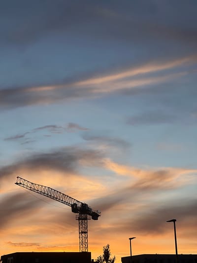 This media file depicts a construction crane silhouetted against a vibrant sunset sky. The sky is a canvas of warm colors, with streaks of orange, yellow, and pink intermingling with softer blues and grays, suggesting a dramatic weather phenomenon or the transition from day to night. The crane, a prominent object in the scene, is a dark, intricate structure with its long boom extending diagonally across the frame.
To the right of the crane, the dark silhouette of a building is visible, with a few windows and architectural details discernible. Two streetlights, also in silhouette, stand tall on the right side of the image. A small tree branch with foliage is visible in the upper right corner, adding a touch of nature to the urban or construction setting. The image captures the industrial presence of the crane juxtaposed with the natural beauty of the sunset.
The time of day is clearly sunset, indicated by the colors of the sky. The weather appears to be clear, allowing for an unobstructed view of the sky's display. While no specific location cues are overtly present beyond the context of "Rotmanka, Poland", the combination of a construction site, buildings, and streetlights suggests an urban or suburban environment. No people are visible, and the overall mood conveyed is one of awe at the natural spectacle and the stark industrial presence. There is no visible text within the image.