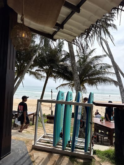 The scene depicts a beachfront area in Dikwella, Sri Lanka, viewed from under a covered establishment. In the immediate foreground, a woven rattan light fixture hangs from a ceiling constructed with burlap-like material and wooden beams. A white security camera is mounted overhead. To the left, a dark wooden vertical pillar or door frame is visible.
In the midground, a male figure wearing a black cap, a black t-shirt with a white graphic, and dark shorts walks from left to right, carrying a white object. He appears to be barefoot or wearing flip-flops. Adjacent to him and slightly right, a white wooden rack holds four turquoise bodyboards, each branded with "JOCK SUPPLIES" and equipped with a yellow coiled leash. Further to the right, a dark wooden table with chairs is positioned. Several indistinct figures are present around this table; one male with lighter hair is partially visible behind the bodyboards, and another male in a dark shirt stands to the right.
The background reveals a sandy beach with numerous tall palm trees. The ocean stretches beyond the beach, exhibiting gentle waves. Several small human figures are visible in the distant water. The sky is overcast. The ground transitions from a compacted area to sand, with patches of grass near the establishment.