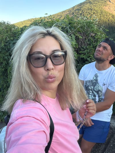 In Partenit, Ukraine, a woman with platinum blonde hair and large dark-rimmed glasses takes a selfie, puckering her lips. She wears a pink crewneck shirt and a silver backpack. Beside her, a man in a white t-shirt with a graphic print and blue shorts looks up and to the right. They are positioned outdoors with lush green foliage and a tree-covered mountainside visible in the background under a clear sky.