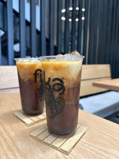 Two plastic cups filled with iced coffee sit on wooden coasters. Cream swirls within the coffee, creating a marbled effect. A black logo, featuring a stylized character, is printed on the cups. The cups are positioned side-by-side, with a portion of each cup visible. The background includes a blurred view of a window with horizontal slats and a light wooden surface.
