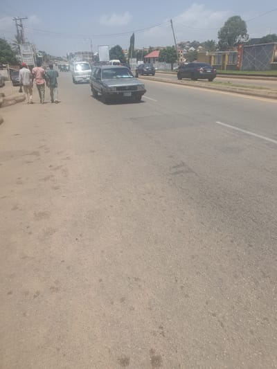 In New Stadium, Nigeria, a street scene unfolds under a bright, hazy sky. A grey Volkswagen Golf sedan is positioned on the right lane of a paved road. To the left of the road, three men are walking on a concrete sidewalk. The man in the front wears a white long-sleeved shirt and dark trousers, carrying a stick. The middle man is in a pink t-shirt and light green trousers, and the third man is in a green shirt and light-colored trousers. Further down the road, various other vehicles are visible, including a white mini-bus and a dark-colored sedan. Buildings line the right side of the road, with some featuring signs and architectural details. The environment is typical of a developing urban area, with overhead power lines and streetlights.