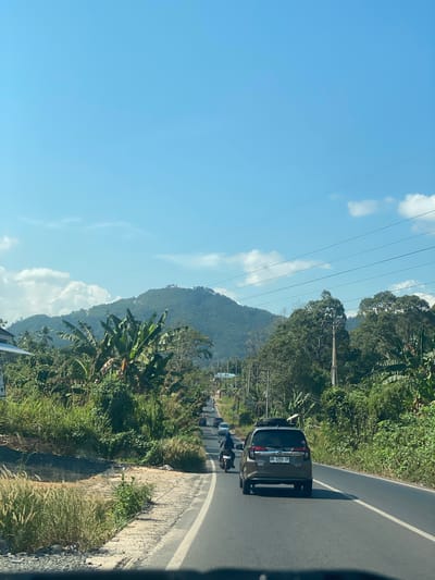 The image depicts a road in the city of Padang Panjang, Indonesia, surrounded by lush green vegetation and rolling hills. The sky is a clear blue with scattered white clouds. In the foreground, a paved road with a white line marking the lanes is visible, with a gray car driving away from the camera. To the left of the road, there is a patch of dry grass and some foliage, while to the right, dense green trees and banana plants line the roadside. In the distance, a mountain covered in trees can be seen. Several other vehicles, including a motorcycle with a rider, are also on the road. Power lines stretch across the sky, supported by utility poles. The scene appears to be a rural or semi-rural area within Padang Panjang.