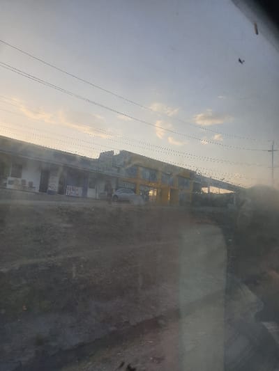 An outdoor shot depicts buildings under a cloudy sky, viewed from behind a dirty glass surface. Power lines are visible against the sky. The right side of the image is dominated by the obscured glass, which reduces clarity and distorts the distant scene. The building facades display a variety of colors and details. The composition emphasizes the separation between the viewer and the external environment.