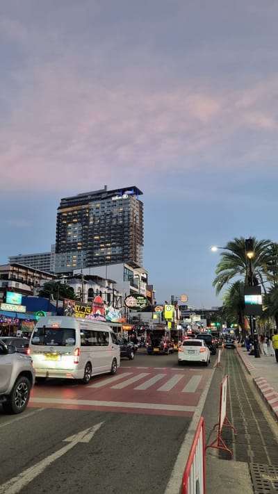 The image depicts a busy street scene in Pattaya City, Thailand, at dusk. A tall Hilton hotel dominates the skyline, its facade illuminated by numerous windows. Below the hotel, a vibrant street is lined with businesses, characterized by colorful neon signs, including "Kamikaze" and "Black & Pearl." The street is congested with traffic, featuring a variety of vehicles such as cars, vans, and motorbikes. Pedestrians can be seen on the sidewalks, and palm trees line the right side of the street. A pedestrian crossing with red and white markings is prominent in the foreground, with cars and a white van waiting at the intersection. The sky above displays a soft gradient of pink and blue, indicative of sunset. The overall atmosphere is one of urban activity and commercial vibrancy.
