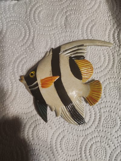 A decorative fish, likely a wall hanging, is placed on a textured white surface, resembling paper towels. The fish is predominantly beige with bold black vertical stripes. It has a yellow eye with a black pupil and a subtle orange-red pectoral fin. Its tail fin and dorsal fin are rendered in yellow with red accents. The background exhibits a repeating pattern of raised circular indentations. The image is taken from a top-down perspective with the fish angled slightly to the left. There is a shadow cast on the lower left portion of the image, suggesting a light source from the top right. No specific location in Gvardeysk, Russia, is discernible from the image.