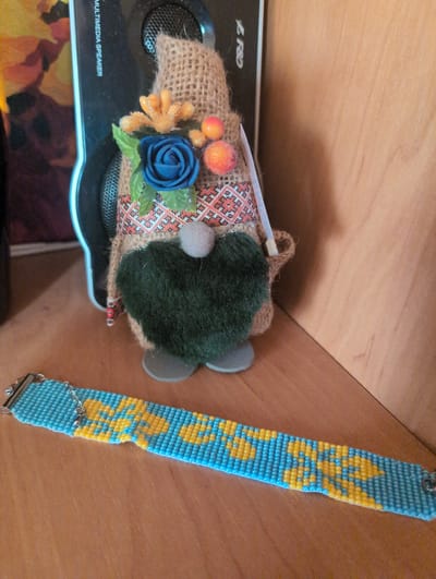 A beaded bracelet with a blue and yellow pattern lies on a wooden surface. Behind it, a gnome-like figurine is propped against a black speaker. The figurine has a burlap body, a dark green fuzzy beard, and is adorned with a blue flower and orange berries. The speaker has the words "MULTIMEDIA SPEAKER" printed vertically on its side. The background appears to be a wooden wall. This scene is located in Kryvyi Rih, Ukraine.