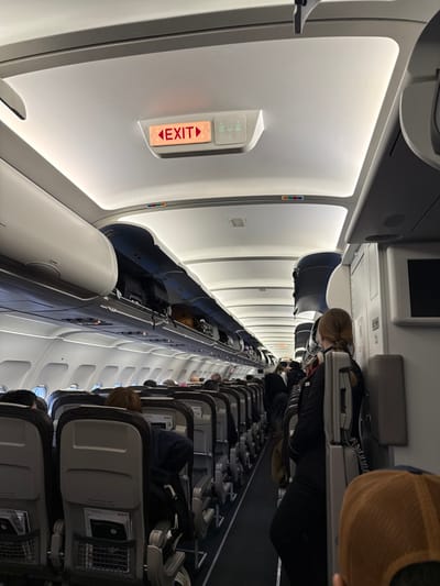 The image depicts the interior of a commercial aircraft cabin, observed from a passenger's perspective, likely at Paris Charles de Gaulle Airport (CDG) in Roissy-en-France, France. The scene shows multiple rows of economy-class seating extending forward down a central aisle. Overhead bins line both sides of the cabin; some on the left are open, revealing luggage stowed inside.
Above the aisle, a rectangular illuminated "EXIT" sign with a left-pointing arrow is visible, alongside a pictogram indicating a lavatory. The cabin's ceiling is white, illuminated by overhead lighting. Numerous passengers are seated in the visible rows, facing forward. On the right side of the aisle, a female flight attendant with light hair in a braid is standing, wearing a dark uniform, and leaning against a cabin partition or wall. She appears to be holding a white rectangular object, possibly a tablet or service item. In the extreme foreground, the back of a person's head wearing a brown baseball cap is visible, indicating the camera's position. The environment suggests either boarding, deboarding, or a pre-flight/post-flight phase of an operational flight.