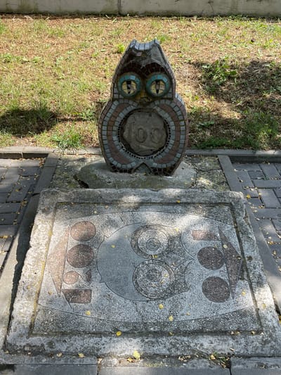 This media file depicts an outdoor scene featuring a mosaic sculpture and a stone carving. The sculpture, shaped like an animal's head with colorful mosaic tiles forming its eyes and body, is positioned to the left of the frame. To the right, a large, rough-hewn stone slab is carved with a stylized owl motif. The owl design includes large, concentric circles for eyes and a triangular head, all set within a stone frame.
The ground surrounding the sculpture and carving is a mix of short, dry grass and a paved walkway made of dark grey interlocking bricks. Scattered yellow leaves are visible on the grass and the paving stones, suggesting it might be autumn. The lighting suggests it is daytime, with shadows cast by the objects. The scene appears to be in a public outdoor space, possibly a park or garden, given the presence of both grass and paved pathways. There is no visible text in the image. The overall impression is one of a quiet, perhaps slightly weathered, outdoor art installation.