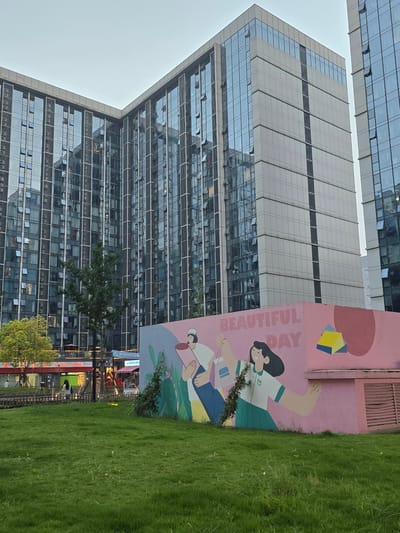 The image depicts a modern urban scene in Linping District, China, dominated by a large, glass-fronted building. The building has numerous windows, reflecting the overcast sky and surrounding environment. In front of the building, a vibrant mural adorns a pink wall. The mural features stylized illustrations of two people, one with dark hair wearing a green and white shirt and a pleated skirt, and another person with a red bandana, holding a book. The text "BEAUTIFUL DAY" is prominently displayed on the mural. Adjacent to the mural is a well-maintained green lawn. The scene suggests a pleasant, possibly early morning or late afternoon, given the soft lighting and the absence of harsh shadows. The overall mood conveyed by the mural is cheerful and optimistic, contrasting with the more imposing architectural structure. A few trees are visible, adding a touch of nature to the urban landscape. Some commercial signage is visible at the base of the building, indicating a public or commercial area. The image is oriented vertically, with the sky at the bottom.