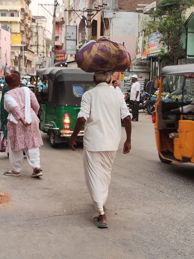 In Puttaparthi, India, a man in white attire carries a large, striped bundle on his head, walking along a street adjacent to auto-rickshaws. Another man, also in white, stands nearby. A woman in a pink and white patterned outfit walks in the same direction. The scene shows a typical urban street setting in India.