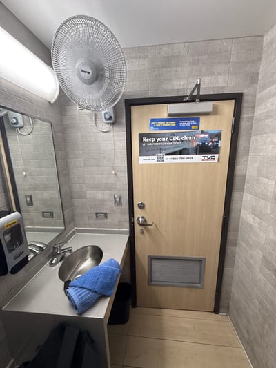 The image shows a restroom with grey tiled walls and a light-colored countertop with a stainless steel sink and faucet. A blue towel is folded on the countertop. To the left of the sink, there is a grey soap dispenser. A large mirror is mounted above the sink. On the wall above the mirror, there is a white oscillating fan.
A wooden door is visible on the right side of the image. The door has a silver door handle, a door closer at the top, and a ventilation grate near the bottom. There are two flyers attached to the upper portion of the door. The top flyer is blue and white and advertises "Love's Freight Factoring & Love's Express Card." The bottom flyer is larger, with a black and white image of a police car with flashing lights, and the text "Keep your CDL clean. Let our pros fight your ticket." It also includes a QR code and a phone number.
The floor is made of light-colored wood planks. In the foreground, a black backpack is visible on the floor. The overall impression is a clean and functional restroom, likely in a public place or truck stop given the nature of the advertisements. The location is not specified within the image itself.