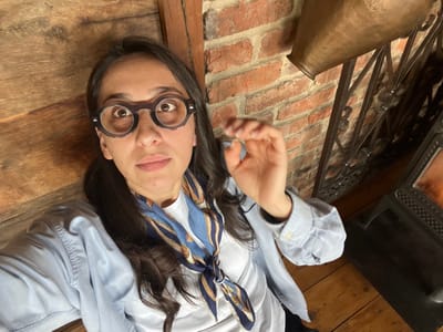 An adult female is depicted in a close-up, upward-angled selfie within an indoor setting. The subject has dark hair, a neutral facial expression, and is wearing round, dark-rimmed eyeglasses, a light blue collared shirt over a white top, and a blue and gold patterned scarf. Her left hand is raised, fingers slightly bent.
The background consists of a vertical wooden wall or paneling on the left, and a reddish-brown brick wall on the right. Between these, and extending into the foreground on the right, is a dark metal decorative railing. Above the railing, a large, dull brass-colored metal vessel is visible against the brick. Below the railing, a dark, cast-iron wood-burning stove with visible orange flames is partially captured. The floor consists of light brown wooden boards.
The precise location within the city of Argveta, Georgia, cannot be determined from the visual evidence provided. The environment suggests a room with rustic architectural elements.