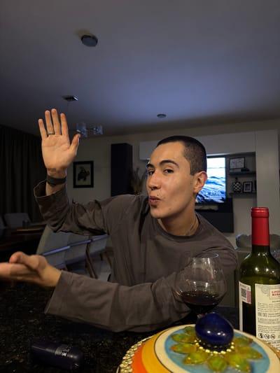 A man is in a room with wine, a wine glass, and decorative objects on a table. The time on a television in the background reads 21:26. The location is indicated by the wine bottle label as Mexico.