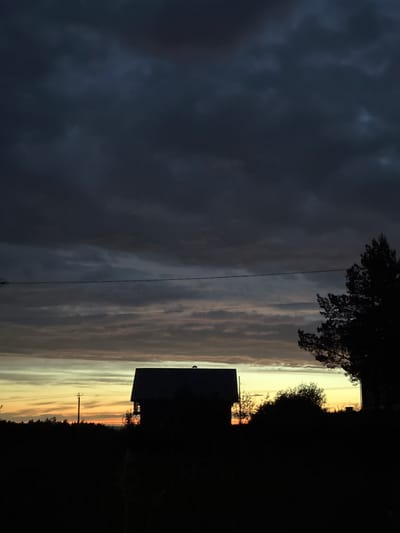 The image depicts a dusk or dawn scene in Прикамский, Russia. A dark silhouette of a house with an adjacent tree occupies the left side of the frame. Behind the house is a band of golden light on the horizon, likely indicating the sun's position. Further to the right, the sky transitions into darker, cloudy formations. A power line stretches across the sky above the house and trees. The environment appears to be a residential area, with a dense, dark background on the left suggesting a forest or wooded area.