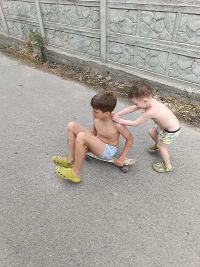 Two young boys are on a paved street next to a concrete fence. The boy on the left is sitting on a skateboard, wearing blue and white patterned swim trunks and bright yellow Croc-style shoes. He has scabs on his knees. The boy on the right is standing and reaching out to touch the shoulder of the boy on the skateboard. He is wearing patterned swim trunks and green Croc-style shoes. The concrete fence behind them has a textured pattern resembling stones and wood grain. Dry weeds and dirt are visible along the base of the fence.